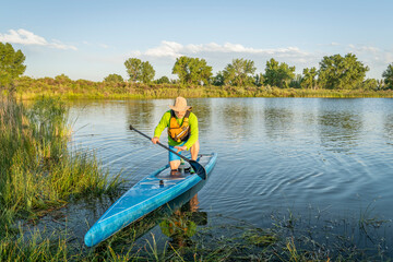 senior male stand up paddler is landing on a grassy lake shore, summer scenery in Colorado