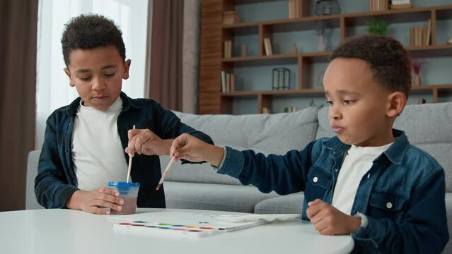 Two little children boys painting at home together. Ethnic brothers African American kids sibling paint picture with acrylic watercolor colors schoolboys indoors enjoy art hobby with paintbrushes