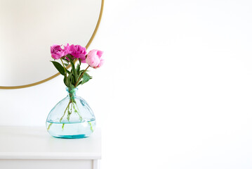 Pink peonies in the apartment. Home decoration, pink peonies on table in white room. Vase with beautiful peony flowers on table in bedroom. Flowers in a vase at home. Mirror reflection, gold frame