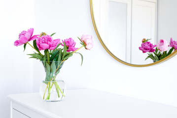 Pink peonies in the apartment. Home decoration, pink peonies on table in white room. Vase with beautiful peony flowers on table in bedroom. Flowers in a vase at home. Mirror reflection, gold frame