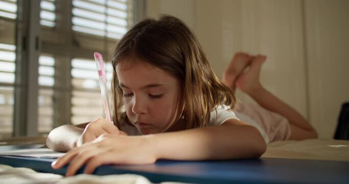 A girl child writes with a pen in a notebook while lying on the floor. A beautiful Caucasian child is studying and doing homework at home. High quality 4k footage