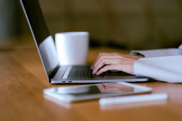 Crop Asian beautiful woman hands typing at laptop on desk with blur tablet, digital pen and a cup of coffee and copy space.