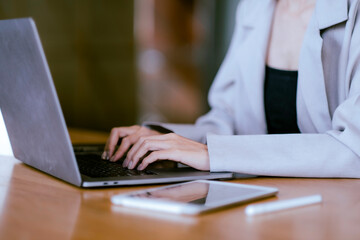 Crop Asian beautiful woman hands typing at laptop on desk with blur tablet, digital pen and with blur gray suit and black dress and copy space.