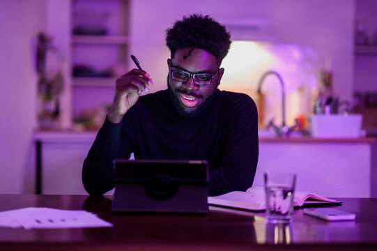 An african american remote worker is sitting at home in a purple lighted room late at night and having an online meeting.