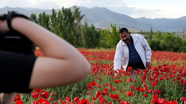 A teenage girl photographs an adult Middle Eastern kind man with a gray beard standing in a field of red poppies in the mountains and picks blooming poppy flowers to make a bouquet and give.