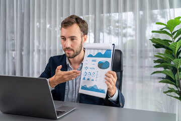 Professional businessman in black suit present financial data or BI paper clipboard to laptop camera during online presentation meeting. Remote work with virtual meeting presentation in office. Entity