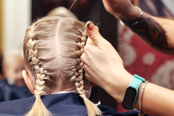 Hairdresser hands makes hairstyle for little blonde girl at barber shop, close-up. Hair stylist braiding braids for pretty kid in barbershop, hair salon. Haircare, beauty concept. Copy ad text space