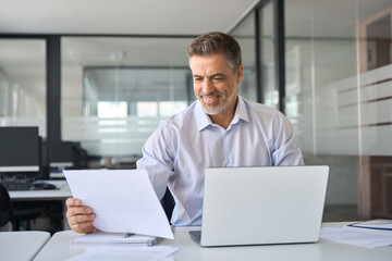 Smiling mature professional business man manager looking at report in office. Happy mid aged businessman company executive working in office with laptop reading financial corporate account documents.