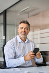 Smiling confident mature business man using phone in office thinking, vertical. Happy mid aged male executive manager holding smartphone using mobile app on cell sitting at work desk looking away.