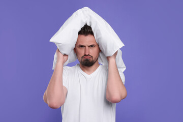 Unhappy man covering ears with pillow on violet background. Insomnia problem
