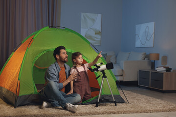 Father and his daughter using telescope to look at stars while sitting in camping tent indoors