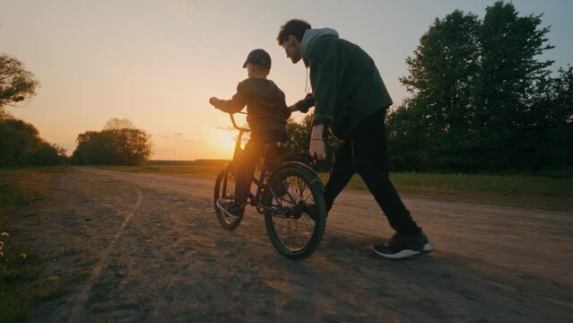 The younger brother learns to ride a bicycle at sunset with the older brother.