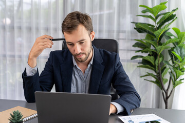 Businessman in black suit working on laptop at his workspace desk. Smart executive researching financial data and planning marketing strategy on corporate laptop at modern workplace. Entity