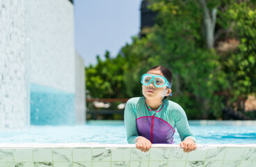 Asian child or kid girl wears swimsuit and swimming goggles on edge swimming pool and happy fun in waterpark for learning swim to exercise on summer school or holiday vacation travel with copy pace