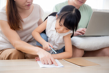 Happy family with mother teaching homework with daughter and father working with laptop on sofa in living room at home, mom explaining schoolwork with kid together, lifestyles and education.