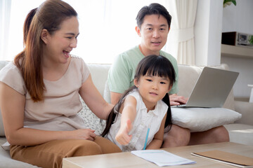 Happy family with mother teaching homework with daughter and father working with laptop on sofa in living room at home, mom explaining schoolwork with kid together, lifestyles and education.