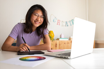Asian young female graphic design student working at home using tablet and laptop looking at camera. Color wheel.