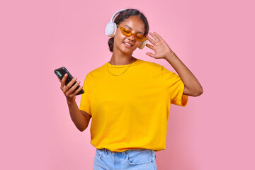 Young serene cheerful Indian woman of adolescence in headphones closing eyes with pleasure listens to music and holds phone for select songs dressed in casual clothes stands on pink background.