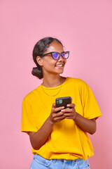 Fototapeta premium Young positive ethnic Indian woman teenager in casual clothes holding phone and laughing chatting with friend from school or arranging walk together on weekend stands on pink background.