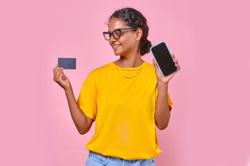 Young happy beauty Indian woman teenager with phone and credit card for payment for internet subscriptions or online services stands on isolated pink background. Smartphone green screen