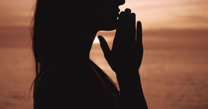 Concept of faith. Close up silhouette of woman folded hands in prayer at orange sunset. Religious woman praying at evening sea. Thoughtful female folding hands into praying gesture.