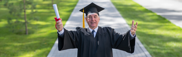 An elderly gray-haired man in a graduate robe spread his arms to the sides and holds a diploma outdoors. Widescreen. 