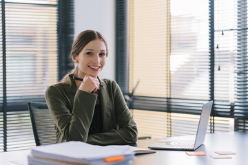A portrait of a millennial businesswoman in a stylish dress is sitting at desk in modern office, using a laptop computer. She appears to be a successful finance manager who is planning work projects.