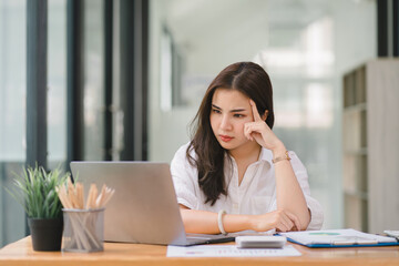 The stressed and exhausted millennial businesswoman is seen sitting at her office desk with her hand on her head, indicating a hard working day where she is overloaded with work.