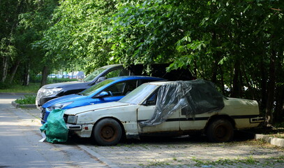 An old broken rusty white car is standing in a parking lot overgrown with trees, Comrade Avenue, St. Petersburg, Russia, June 20, 2023