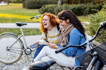 Two girl friends sitting on bench, looking at mobile phone and smile in public par with bycicle on background