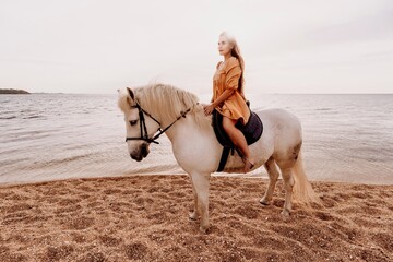 A white horse and a woman in a dress stand on a beach, with the sky and sea creating a picturesque...