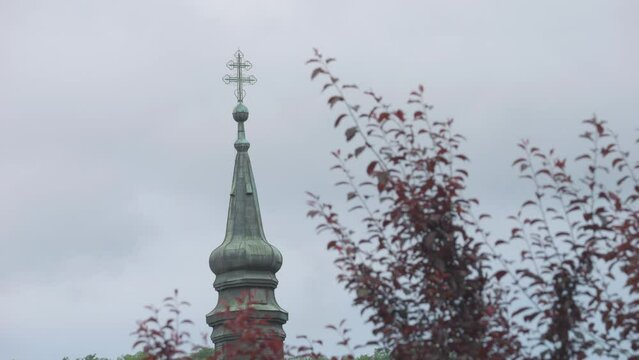Serene footage of a church tower with a cross on top behind branches with leaves on top on a windy day