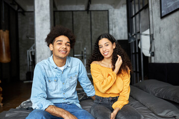 Young african ethnicity couple of 20s posing in front of camera with happy smiling faces, spending weekend together, sitting on gray bed in loft apartment, female touching her hair, feeling shy