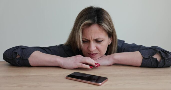 Lonely sad girl is waiting for phone call sitting at table at home. Nervous frustrated woman waiting for a call or sms on smartphone