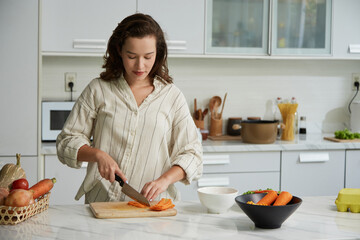 Young woman cutting carrots in matchsticks for salad at kitchen counter