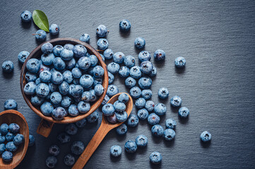 The blueberries in a wooden bowl and spoon and scattered on a black stone surface.