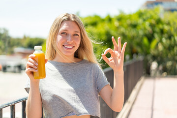 Young blonde woman holding an orange juice at outdoors showing ok sign with fingers