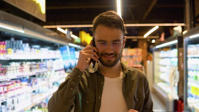Young man walks in a supermarket makes purchases, chooses products and talks on the phone, consults with his wife about the shopping list