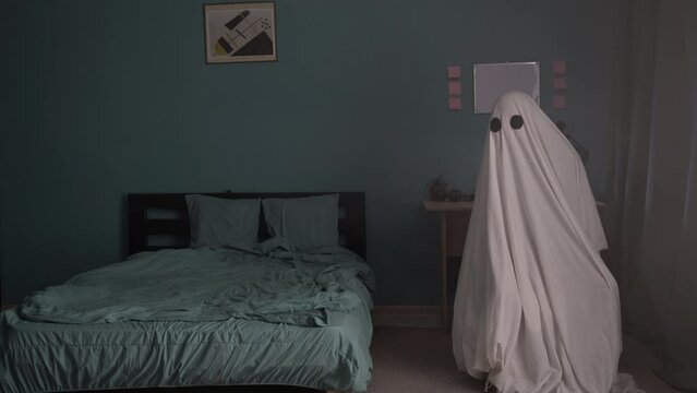 Ghost in a sheet sits in room near bed, holiday of dead