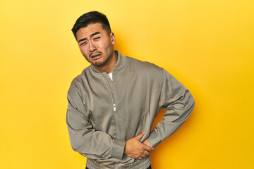 Stylish Asian man in gray jacket on yellow studio having a liver pain, stomach ache.