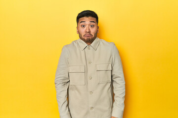 Fototapeta premium Asian man in stylish gray shirt on yellow studio shrugs shoulders and open eyes confused.