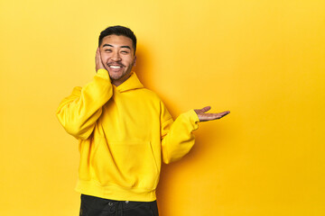 Bold Asian man in yellow hoodie on yellow studio holds copy space on a palm, keep hand over cheek. Amazed and delighted.