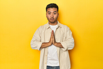 Casual Asian man with open shirt, white tee on yellow studio praying, showing devotion, religious person looking for divine inspiration.