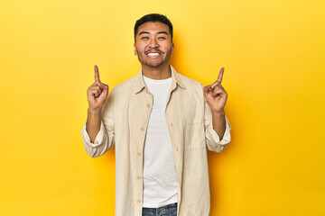 Casual Asian man with open shirt, white tee on yellow studio indicates with both fore fingers up...