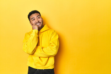 Bold Asian man in yellow hoodie on yellow studio relaxed thinking about something looking at a copy space.