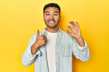 Asian man with invisible dental corrector, yellow studio backdrop smiling and raising thumb up - obrazy, fototapety, plakaty