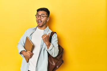 Asian student with backpack and notebook, yellow studio backdrop points with thumb finger away, laughing and carefree.