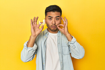 Asian man with invisible dental corrector, yellow studio backdrop with fingers on lips keeping a secret.