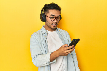 Asian man making a playlist on his phone while listening to music, on yellow background.