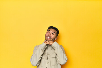 Obraz premium Asian man in stylish gray shirt on yellow studio keeps hands under chin, is looking happily aside.